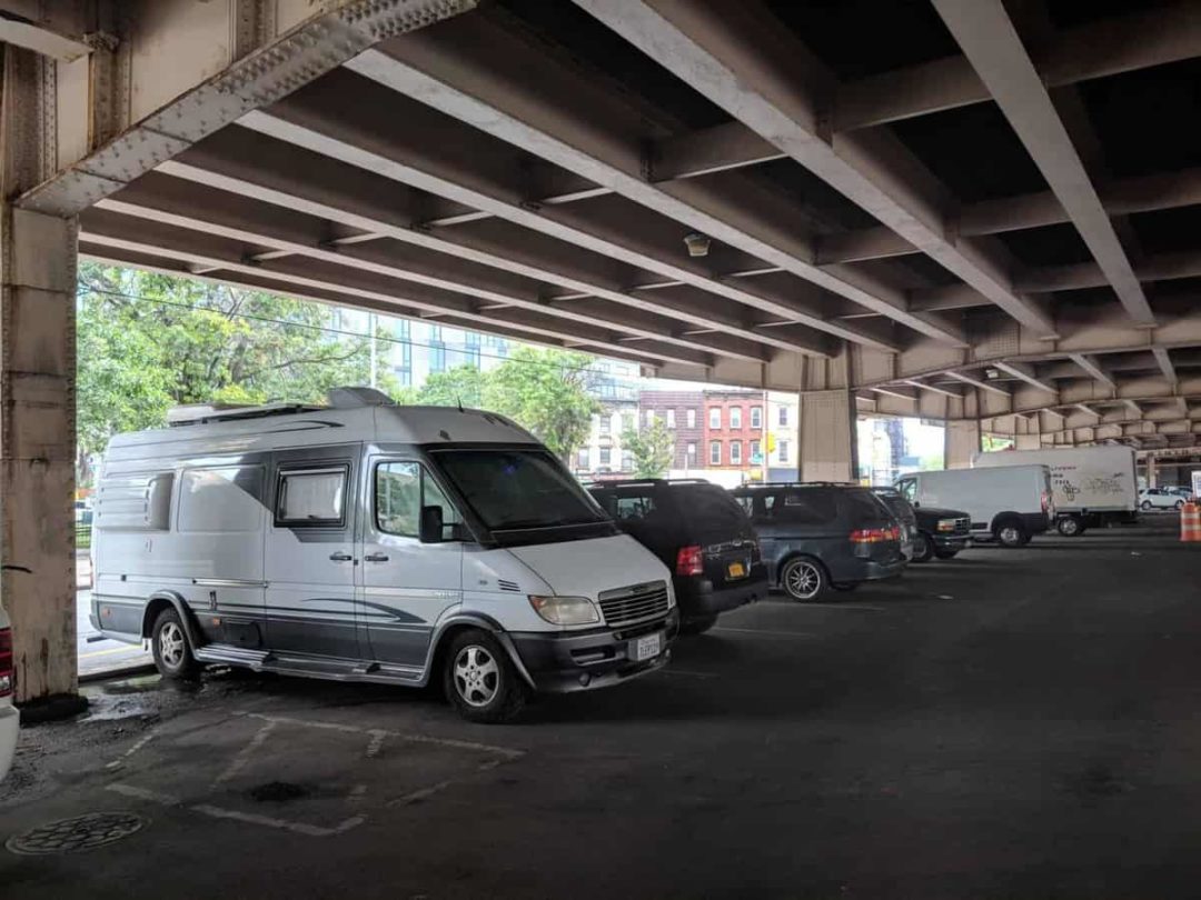 A lineup of vans and cars parked under a freeway overpass