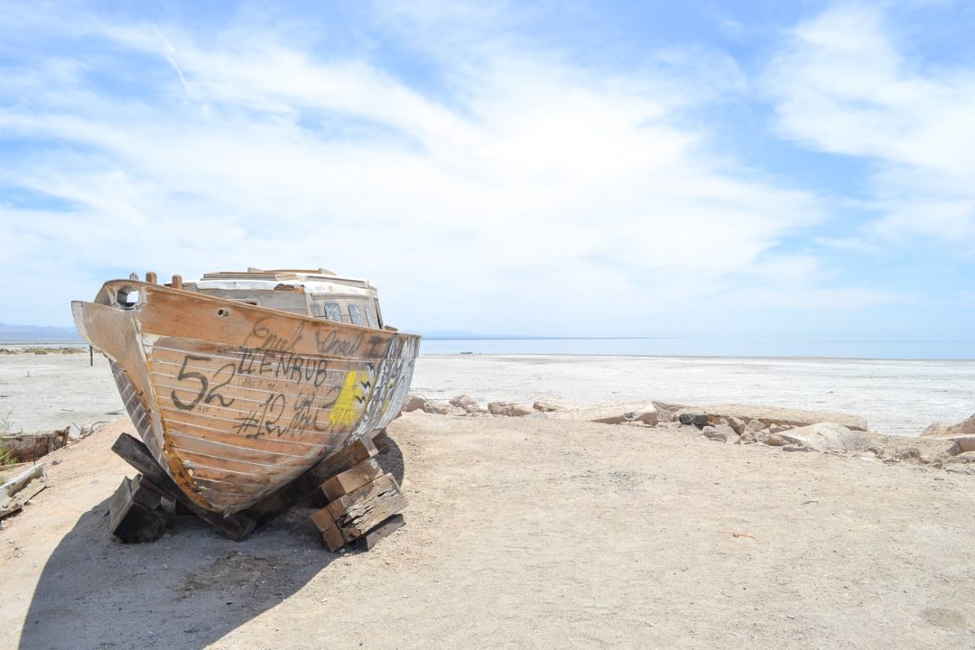 An old wooden boat covered in grafitti sits on a sandy shore with a blue lake in the background