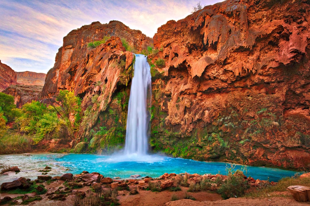 a waterfall off a red rock cliff plunges into a blue pool