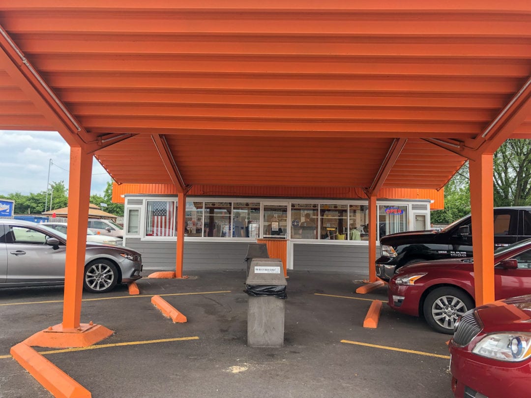 The drive-in at The Root Beer Stand is an orange shelter leading into a grey building with a storefront full of windows.