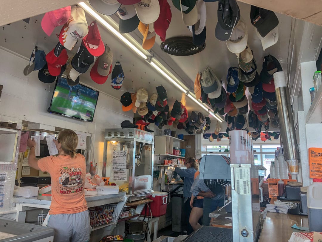 Employees work to fill orders behind a bar counter. Above them, dozens of hats of all colors hang from the ceiling. Popcorn pops in the background.