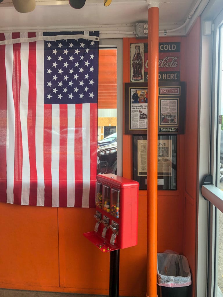 An American flag and gum ball machine sit in front of a bright orange wall in The Root Beer Stand.