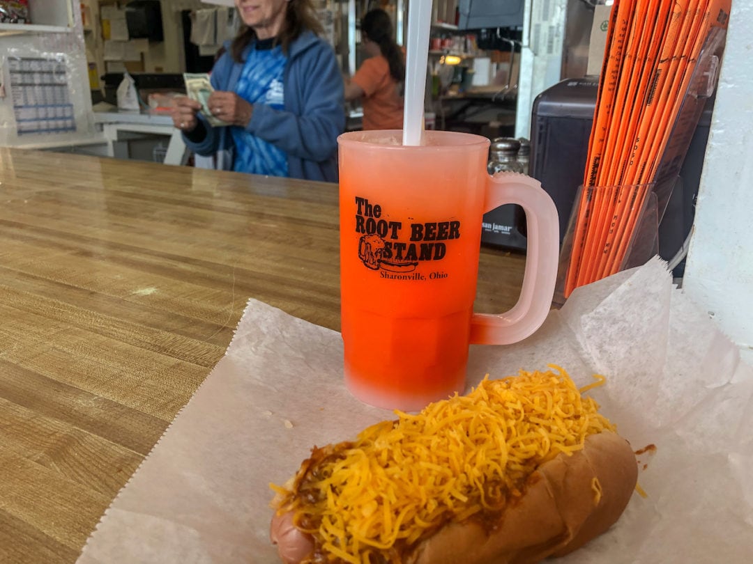 A root beer float in an orange souvenir mug that reads "The Root Beer Stand, Sharonville, Ohio," next to a six-inch chili cheese dog.