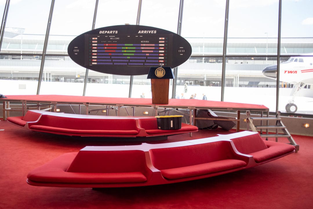 a red sunken lounge with red carpet and a black and white arrival board with glass windows and a plane in the background