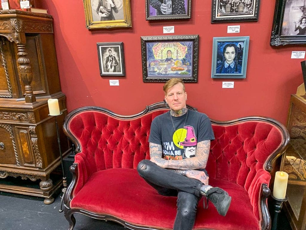 Eric Wessel, owner, sits in a red velvet love seat in front of Adams Family portraits on the wall.