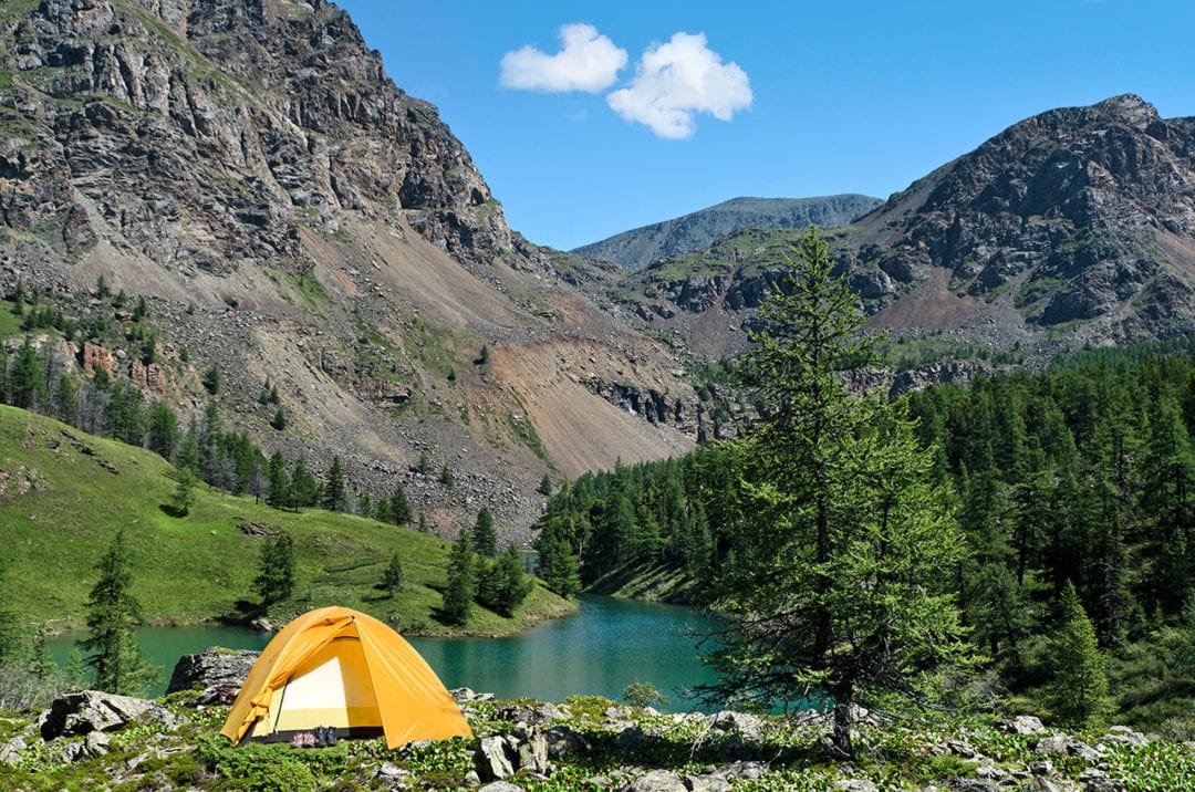 A tent sits next to a small lake surrounded by mountains and forest 