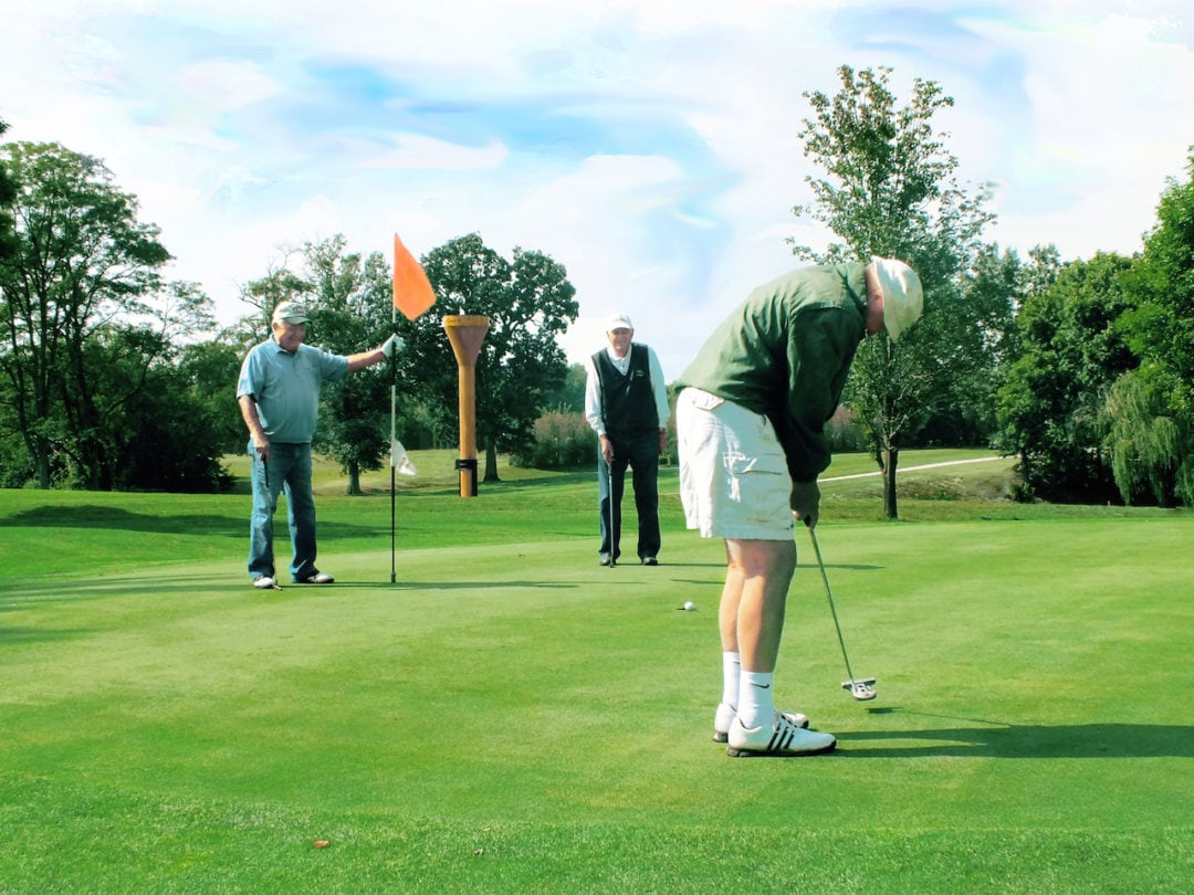 three men putt balls into the hole in front of a giant golf tee