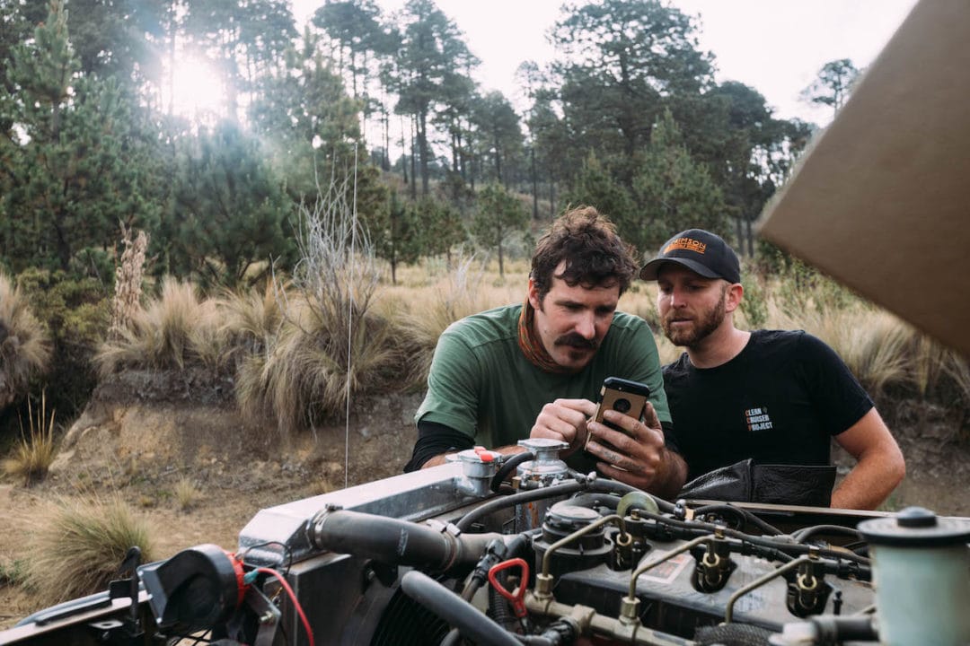 Two men stand in front of an exposed engine, checking their phone