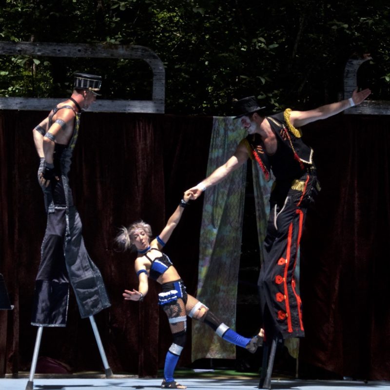 Two men on stilts hold up a circus performer