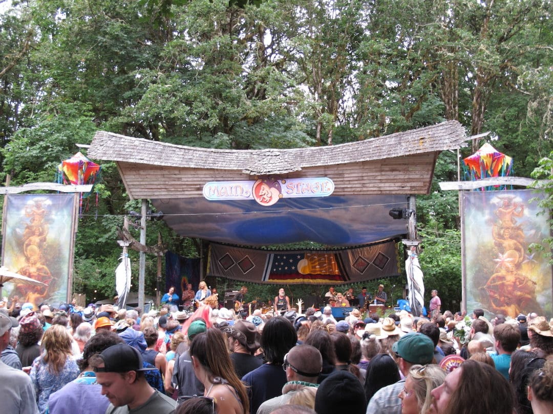 hundreds of people stand in front of a band performing on the main stage, in front of a wooded area