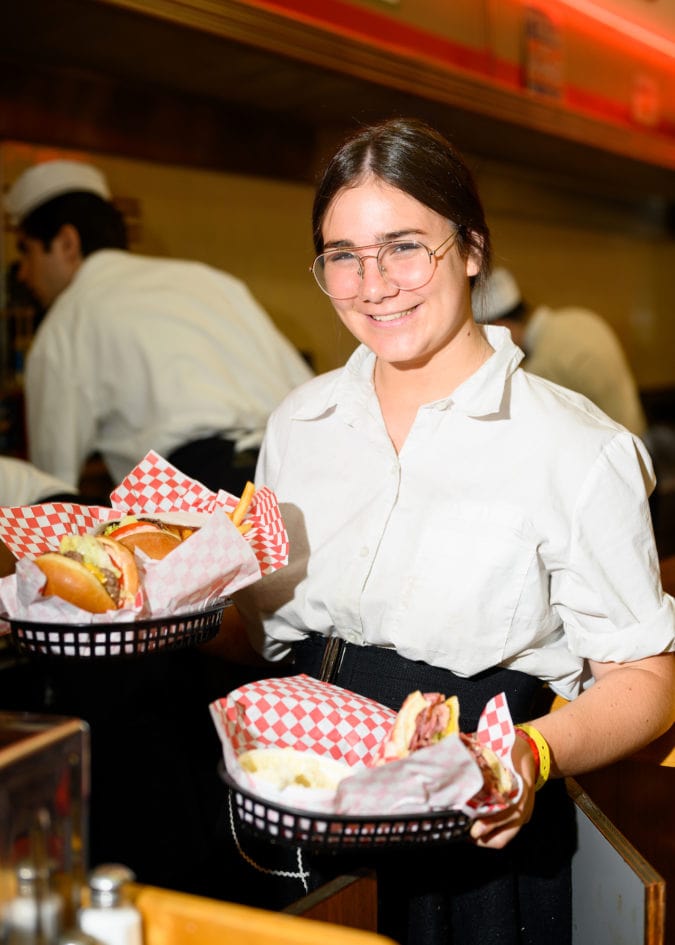 a young girl in glasses holds to baskets filled with burgers
