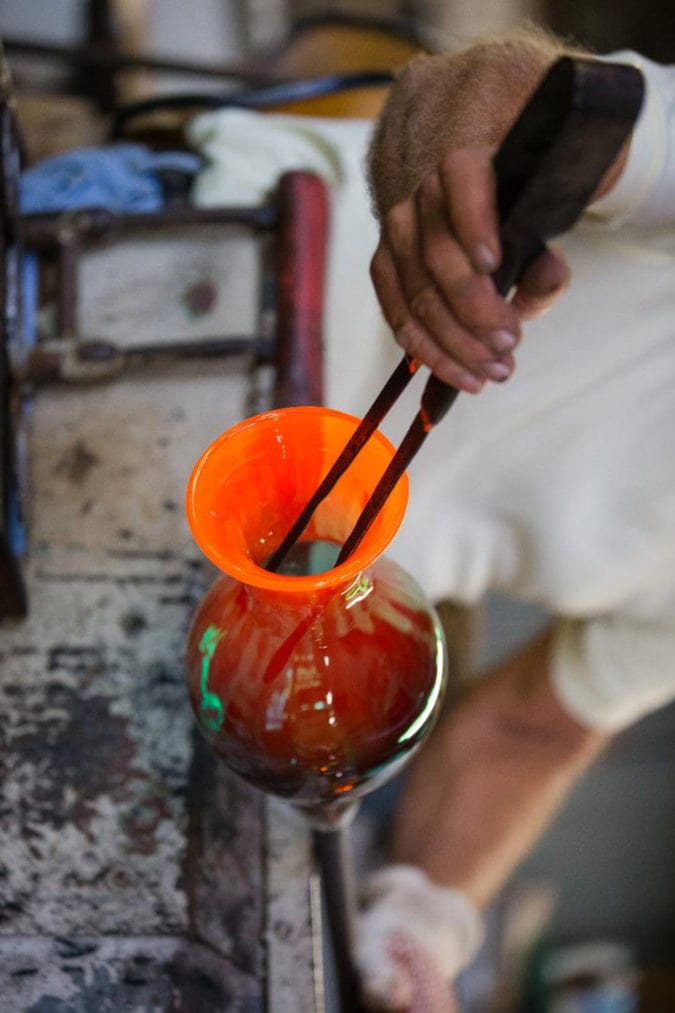 A Blenko artisan uses a tool to give a bright orange vase its final shape.