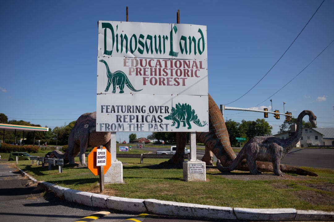 A sign for dinosaur land flanked by several fiberglass dinosaurs on a crossroads in virginia