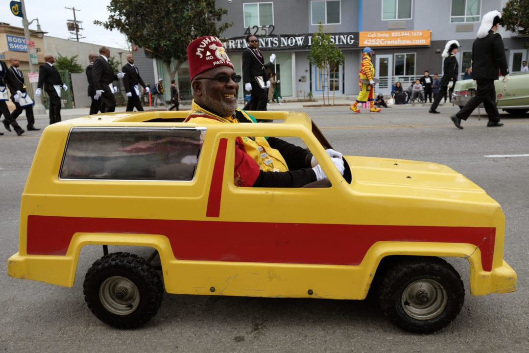 Desert High Priest and Prophet, Bishop Dr. Reginald “Reggie” Black, drives the fun car around in circles, waving and posing with the crowd for photos. Black is a member of the Shrine Mason Fraternity Muharram Temple No. 95 in the Oasis of Santa Monica, California. The brotherhood is an Ancient Egyptian Arabic Order of the Nobles of the Mystic Shrine. The fraternity was born out of the Freemasons, and bases its philosophy on fun, family, fellowship, and philanthropic work.