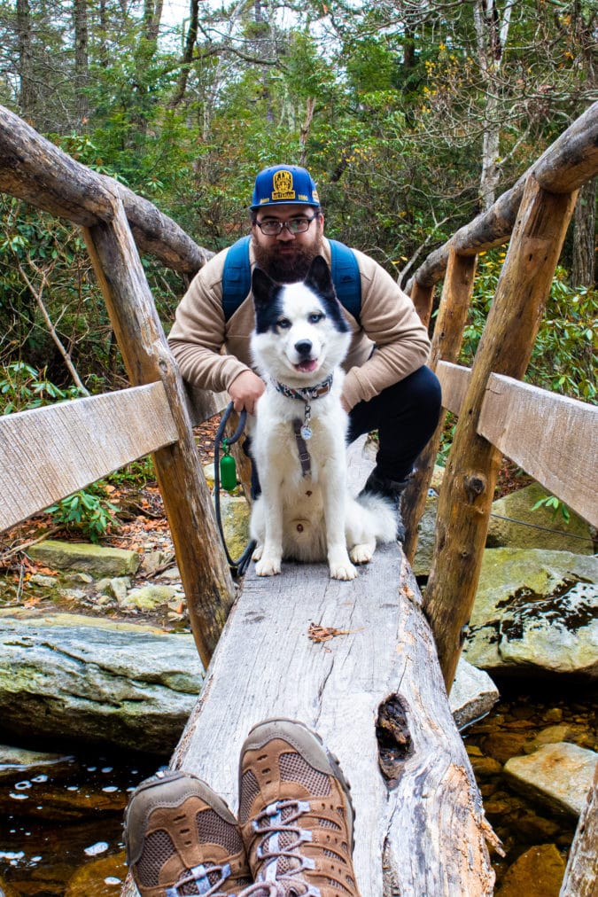 Man with beard, glasses and a hat sits behind and poses with black and white husky on a fallen log bridge