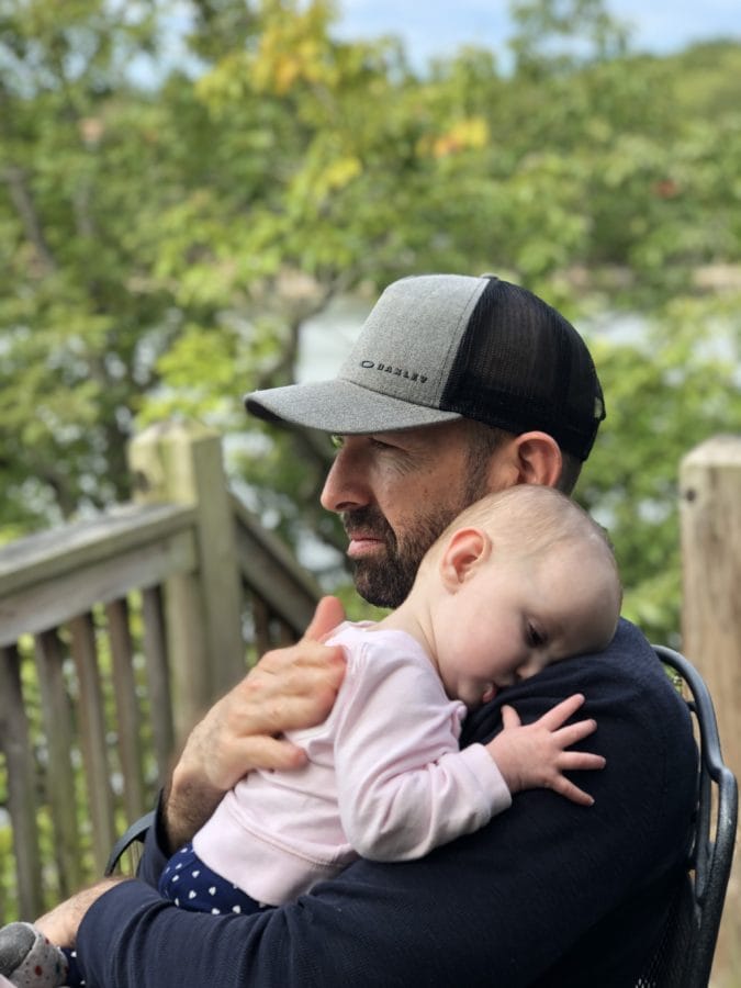 Dad in a hat holding his baby daughter on his shoulder looking out over the water