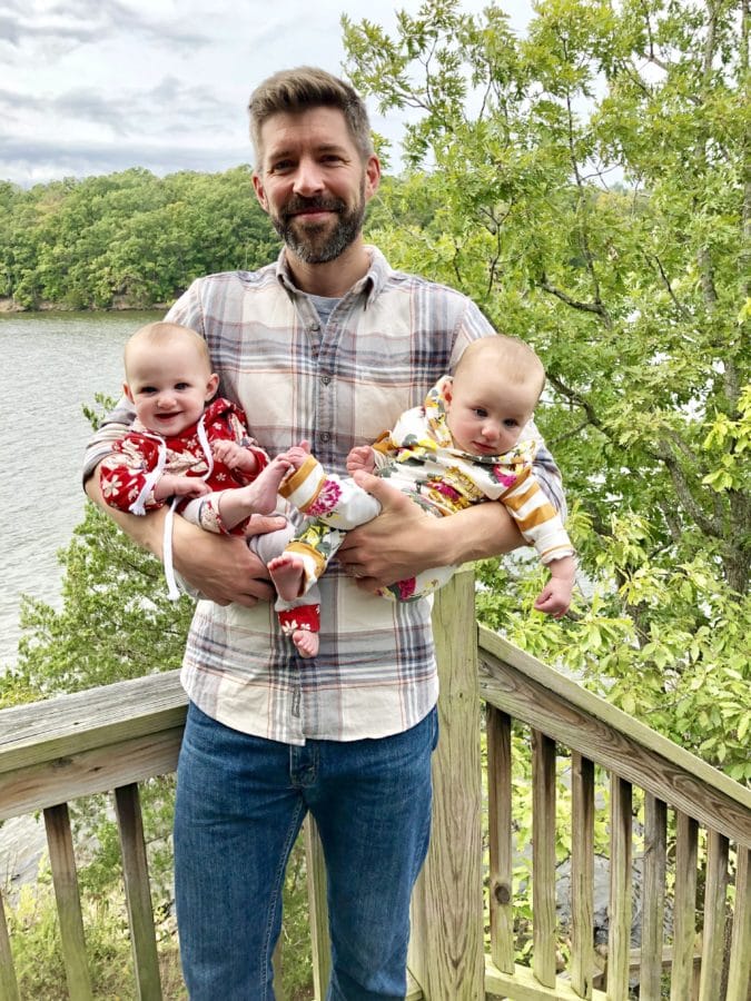 Smiling dad standing on a deck holding twin baby girls
