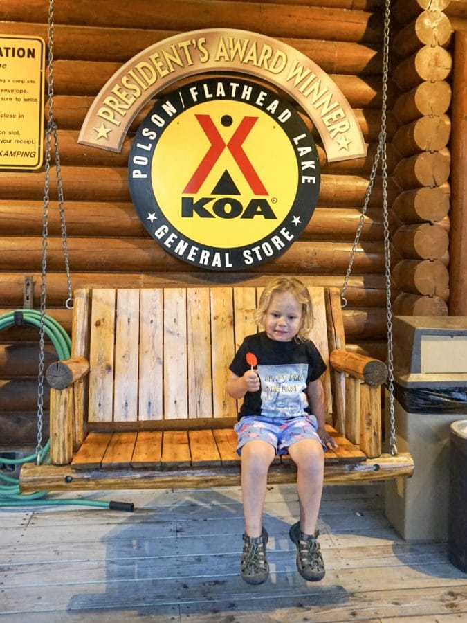 Young boy sits on a swing holding a red lollipop in front of a KOA campground sign