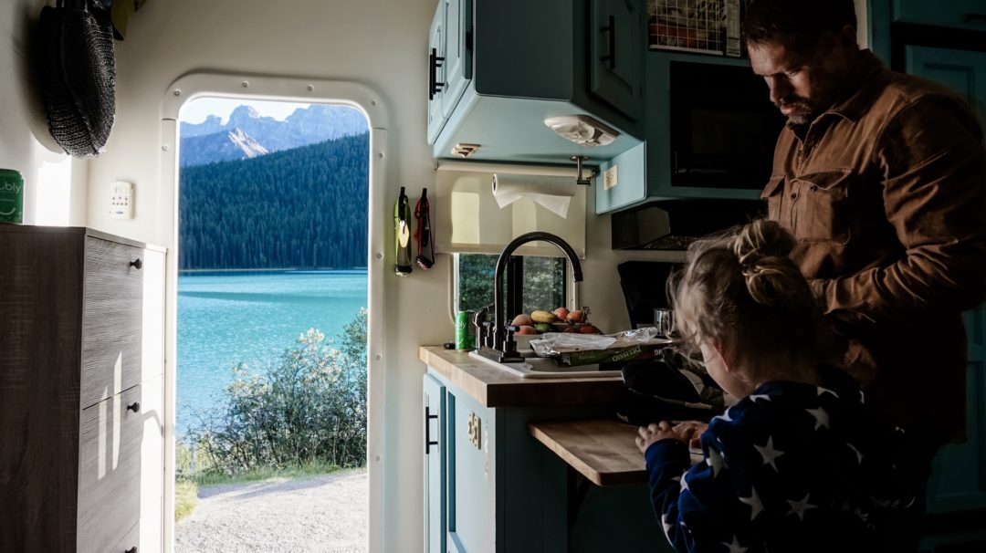 Young boy and father inside an RV in the kitchen with a view of a crystal blue lake out the front door
