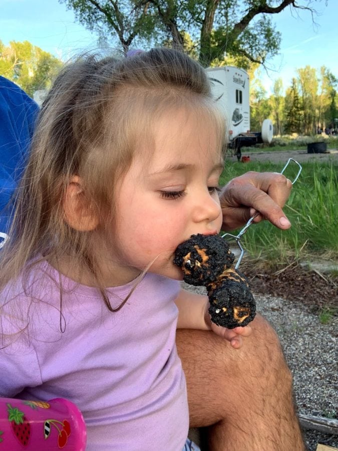 Young toddler in pink shirt takes a bite out of a toasted marshmallow off the skewer