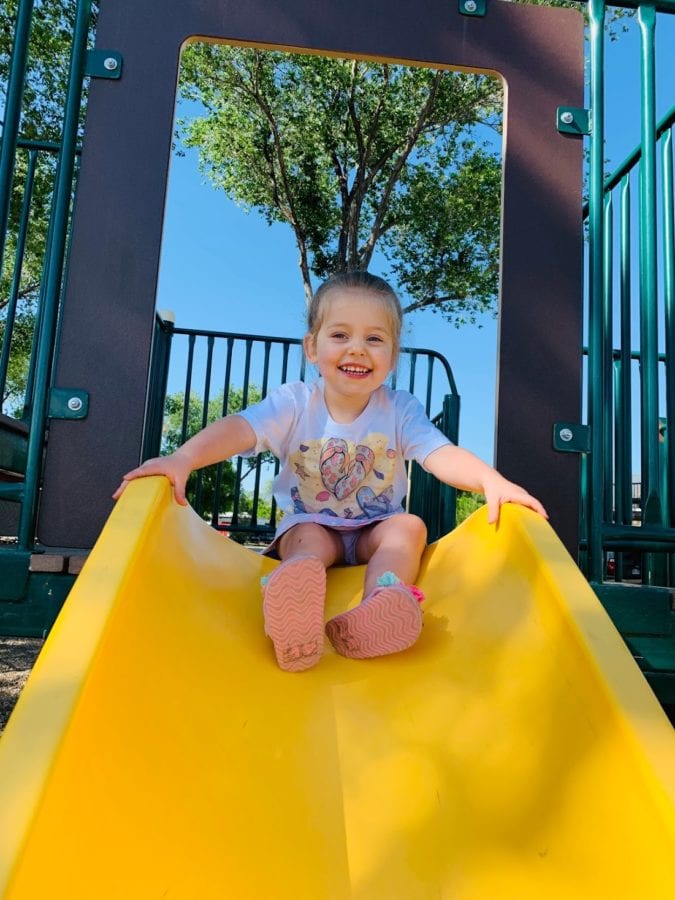 Young toddler sits at the top of a yellow slide smiling down ready to slide down