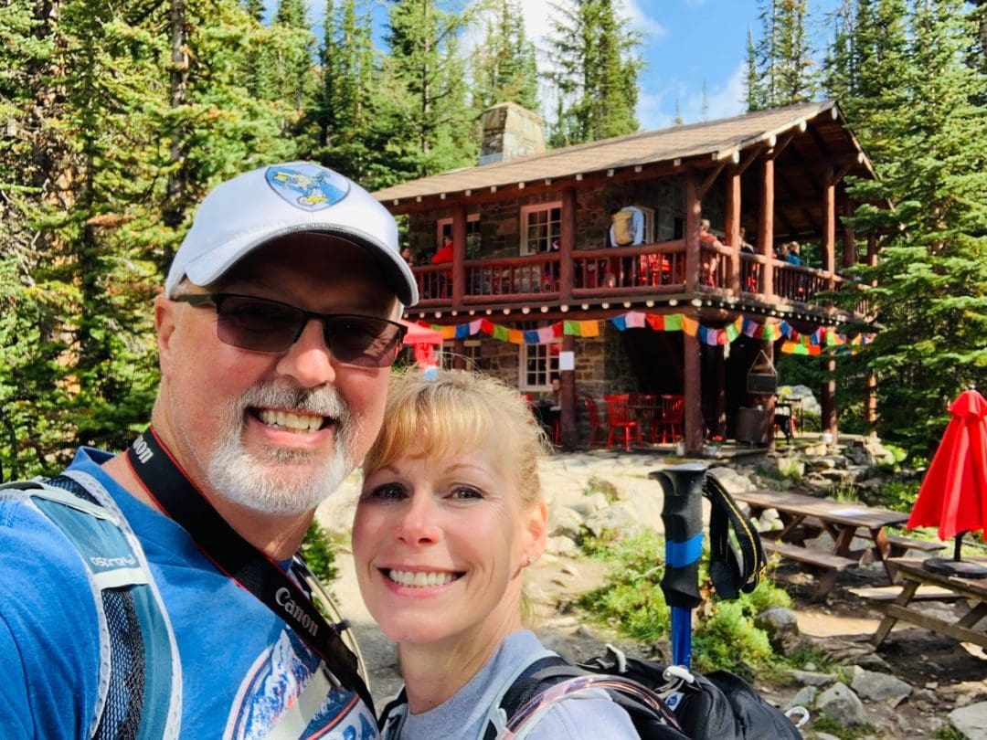 Selfie of hiking couple wearing backpacks in front of old wooden cabin covered in flags and surrounded by pine trees