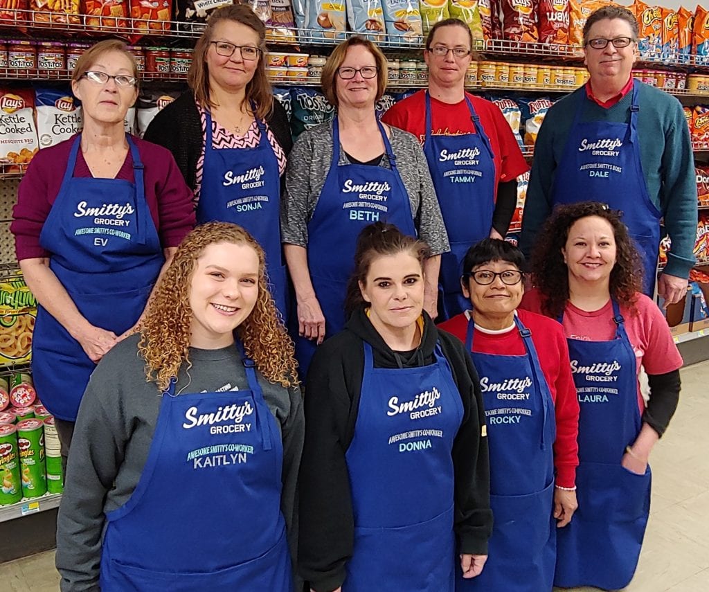 Group of people in two rows standing in a grocery aisle wearing matching blue aprons