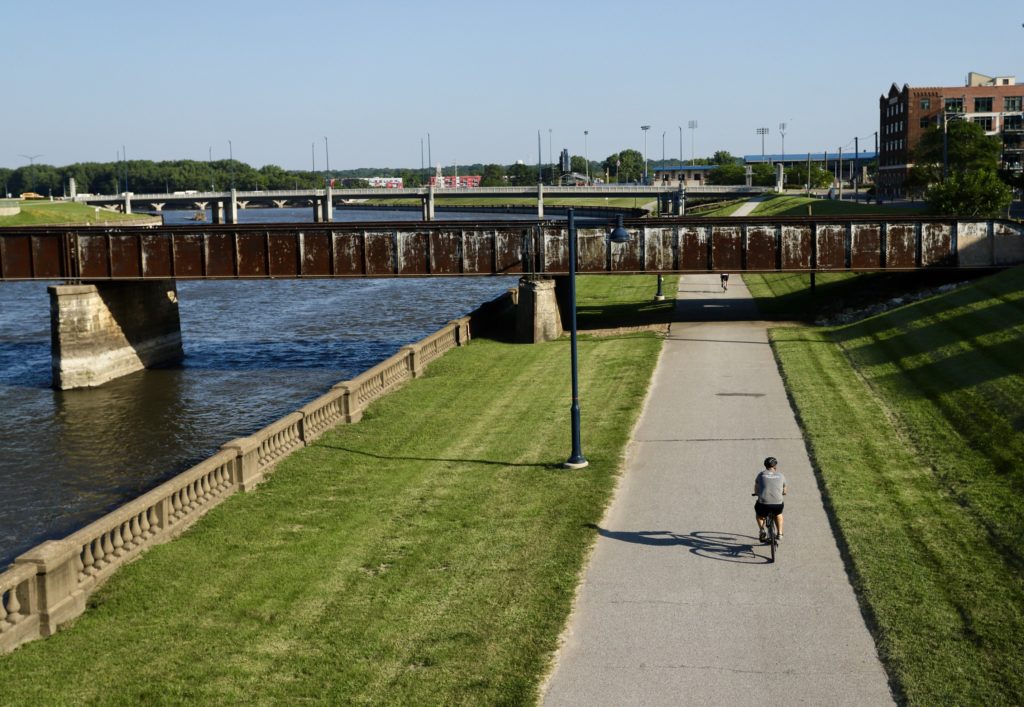 Lone biker on paved path next to Des Moines River and green grass