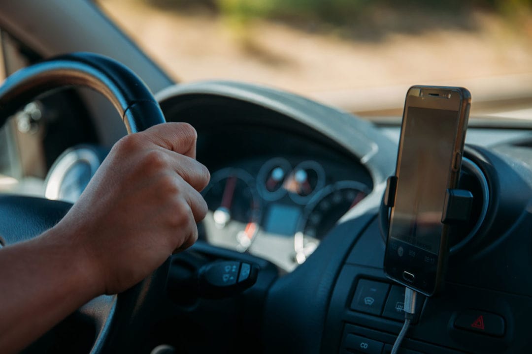Close up of phone holder with smartphone inside a car, one hand on the steering wheel. 