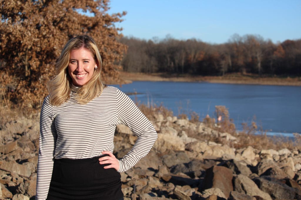 Photo of a blonde woman in a striped shirt standing in front of a rock riverbank