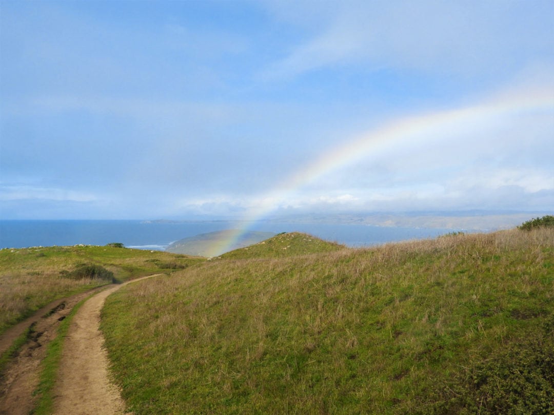 A rainbow above a hiking trail with the Pacific Ocean in the background.