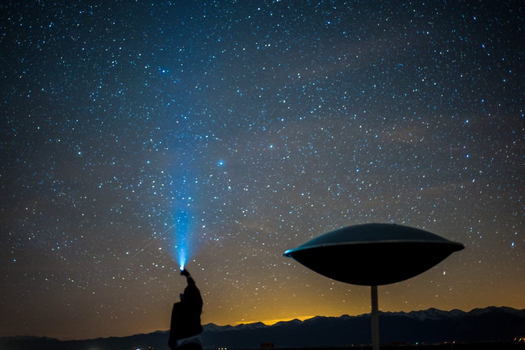 the silhouette of a person shining a flashlight up into a night sky full of stars next to a saucer-shaped object