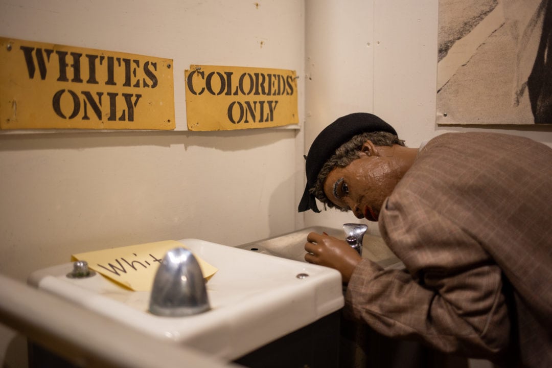 A figure is bent over a water fountain with signs above stating "whites only" and "coloreds only"