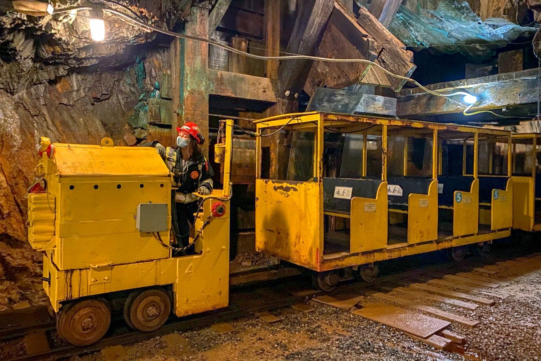 A bright yellow train driven by a woman in a red hard hat inside of an underground mine