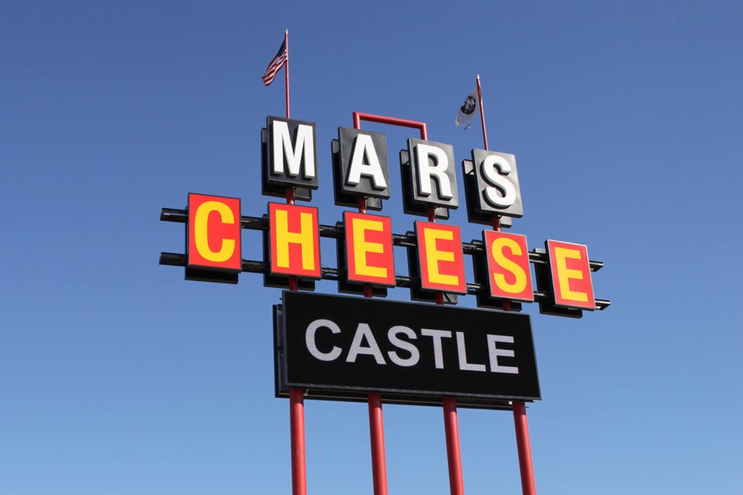 A black, red and yellow sign that spells out "Mars Cheese Castle" topped by two flags
