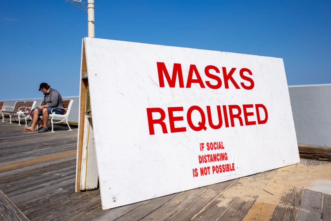a large white sign sits on a boardwalk and says "masks required if social distancing is not possible"