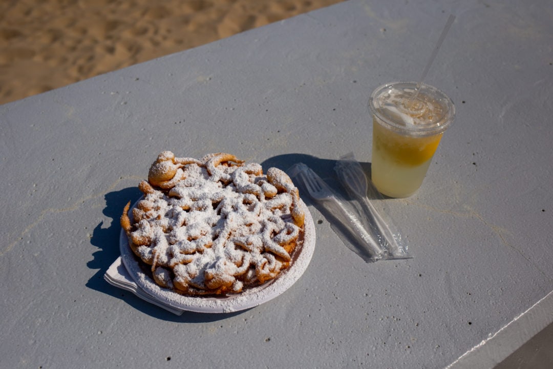 a funnel cake topped with powdered sugar and a cup of lemonade by the beach