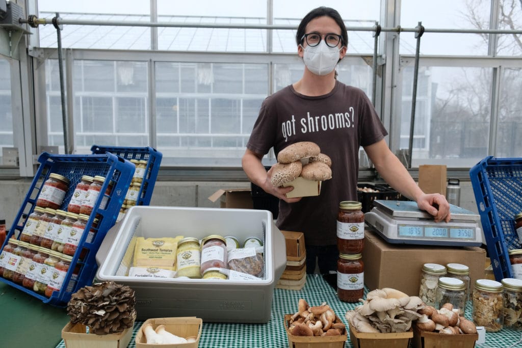 a man wearing a facemask and a brown shirt that says "got shrooms?" holds mushrooms and stands in front of a table with farm products for sale