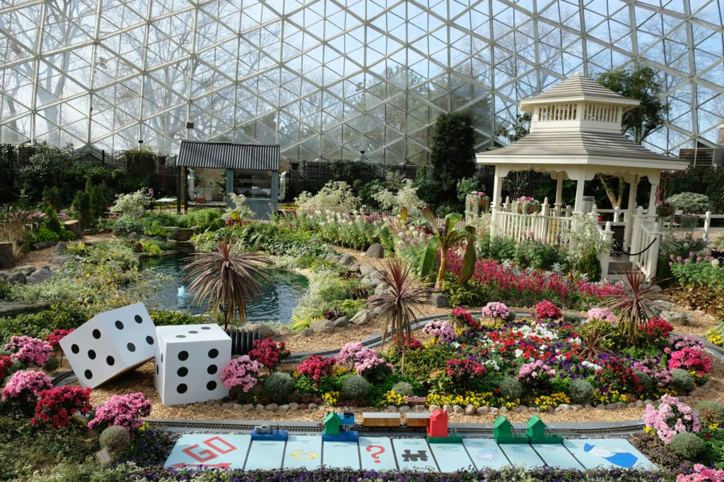 colorful flowers and monopoly game board pieces at a holiday train show inside of a domed glass greenhouse