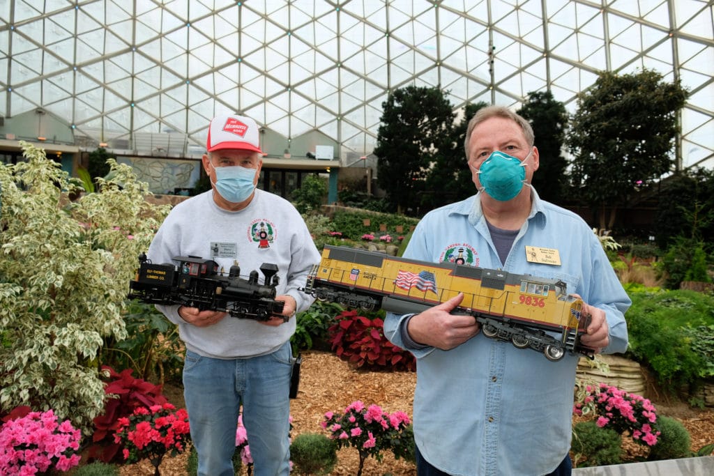 two men wearing face masks hold model trains in front of colorful flowers