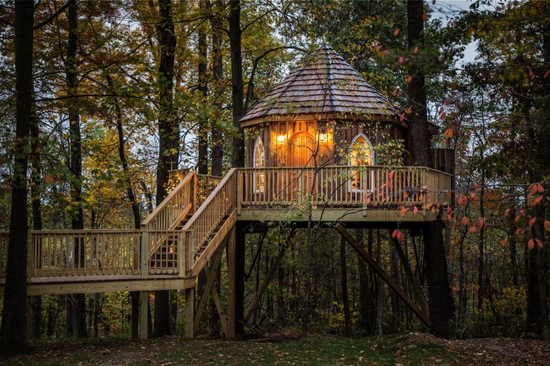 A wooden treehouse set in the woods with a wooden walkway and staircase