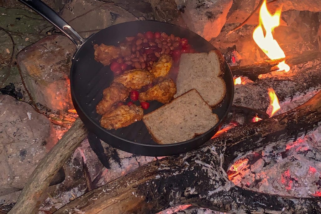 An iron skillet with beans, bread and chicken cooking on a campfire