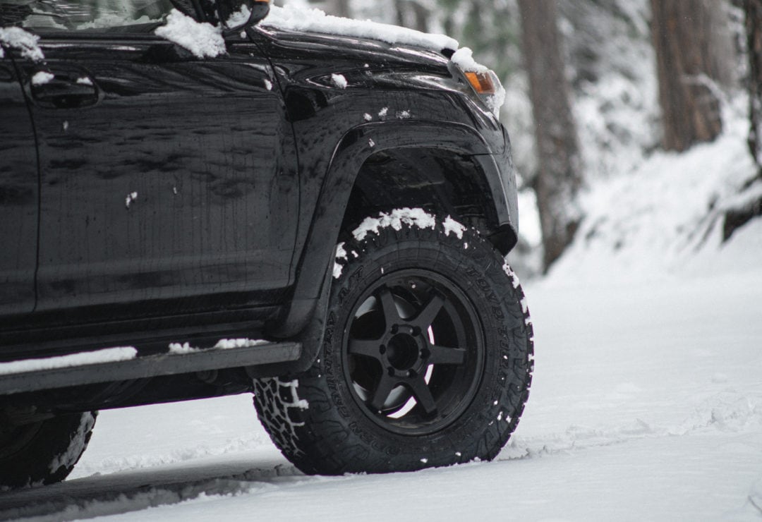 Close up of black Toyo tires with black rims lightly dusted in snow and surrounded by snowy backdrop