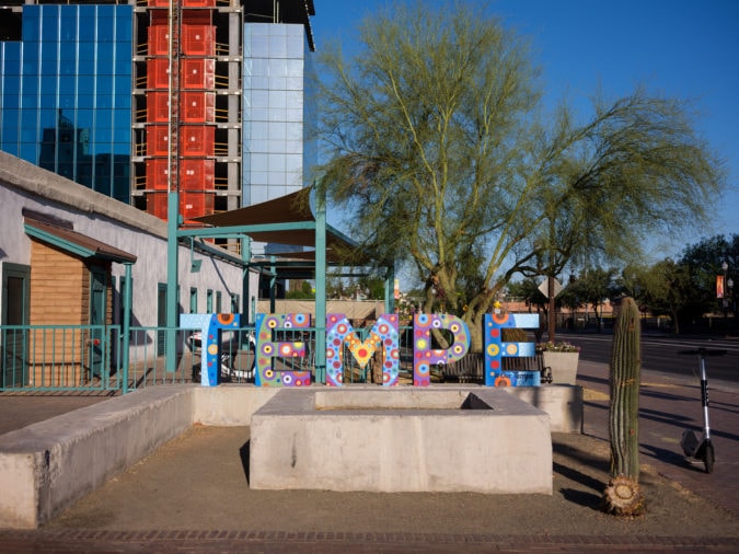 Colorful painted sculpture spelling TEMPE in front of green tree and modern glass building