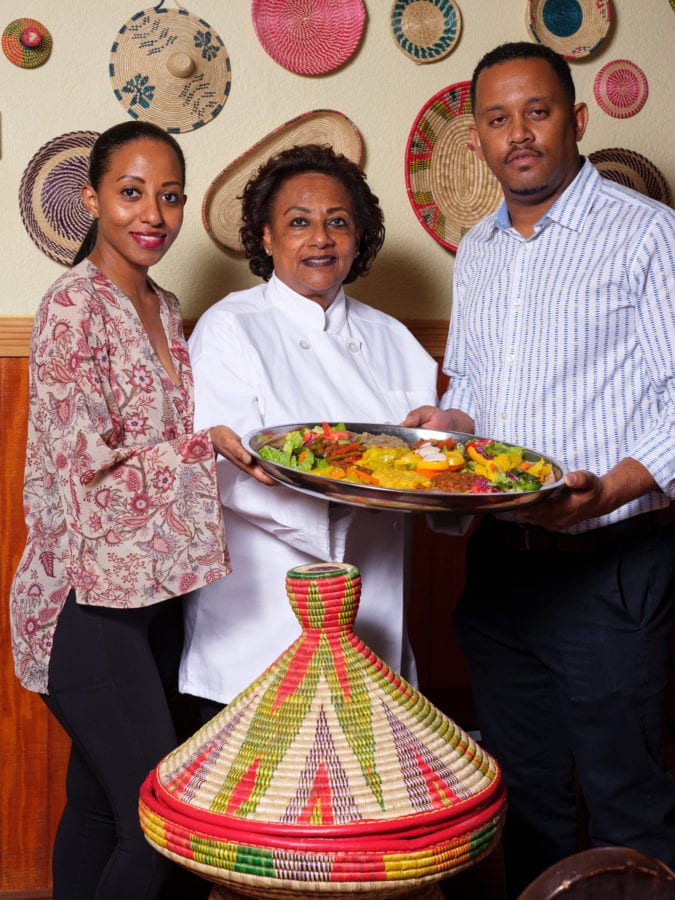 Three people, two women and one man, stand smiling at camera while presenting large silver platter of colorful food