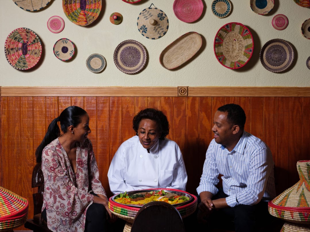 Three people, two women and one man, sitting around a large platter of colorful food with decorative woven baskets on the wall behind them