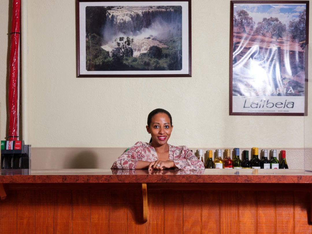 Women stands behind tall wooden counter smiling at camera with bottles of wine lined up behind her