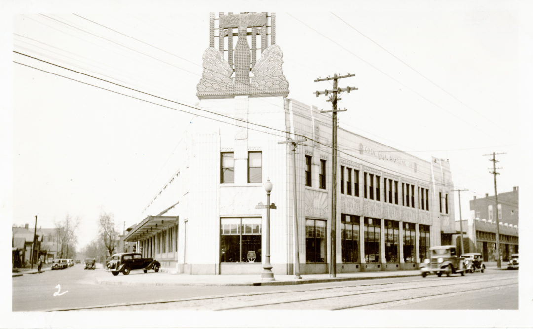 a black and white photo from 1935 of a white art deco coca-cola factory with a sign featuring a soda bottle