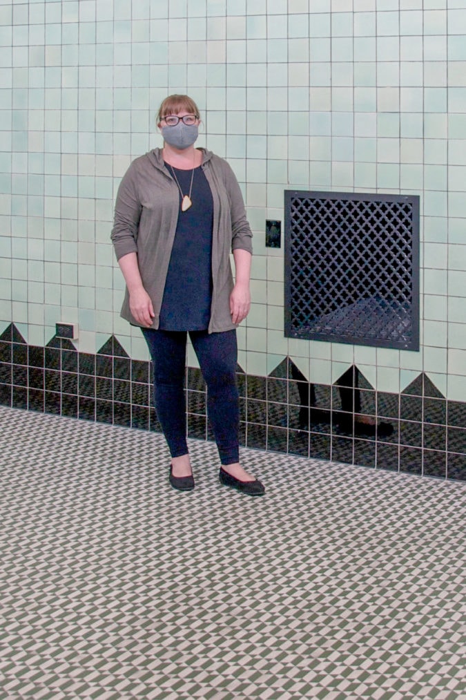 A white woman with reddish blonde hair wears a face mask, jeans, and a sweater and stands in front of a green and black tiled wall