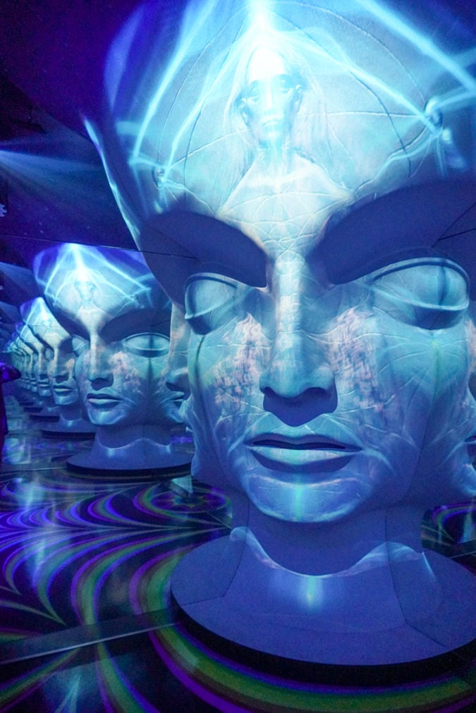 A room with a big human head sculpture surrounded by blue light and mirrors, which create an optical illusion. 